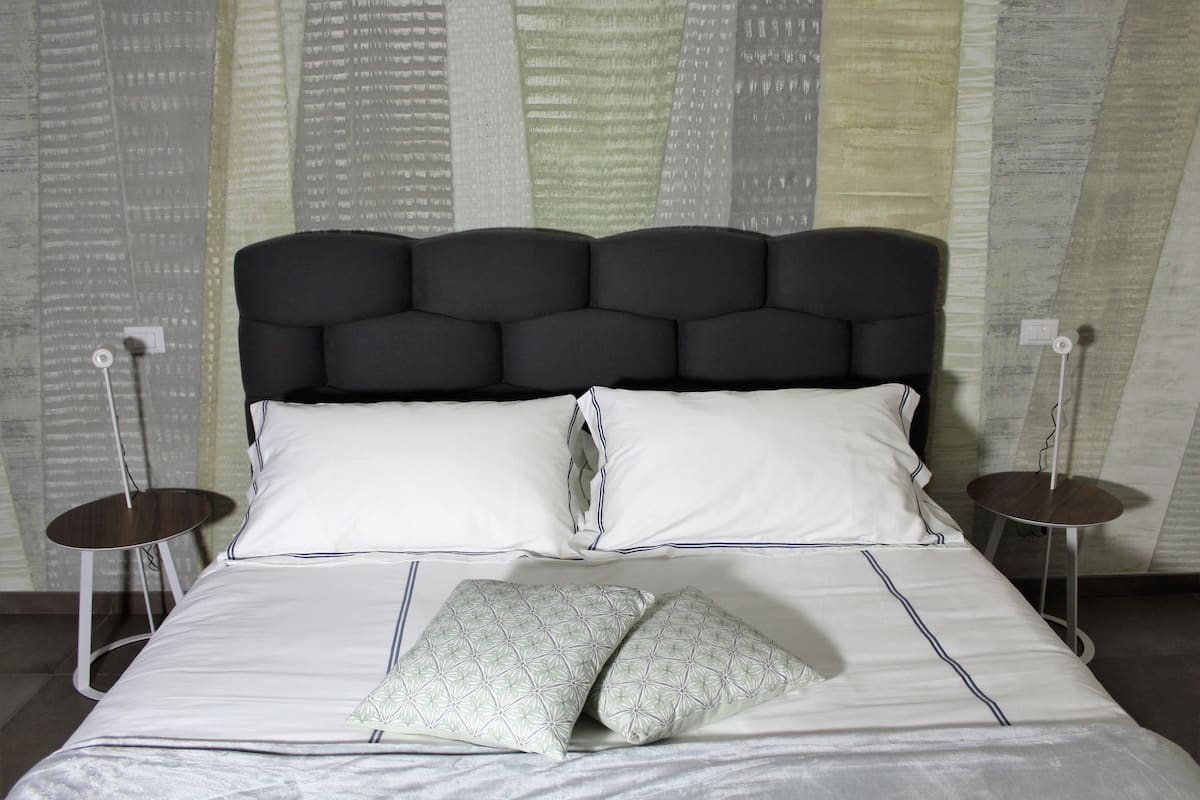 A neatly made bed with a black upholstered headboard, white linens, and two decorative pillows. There are two bedside tables with lamps on either side of the bed.