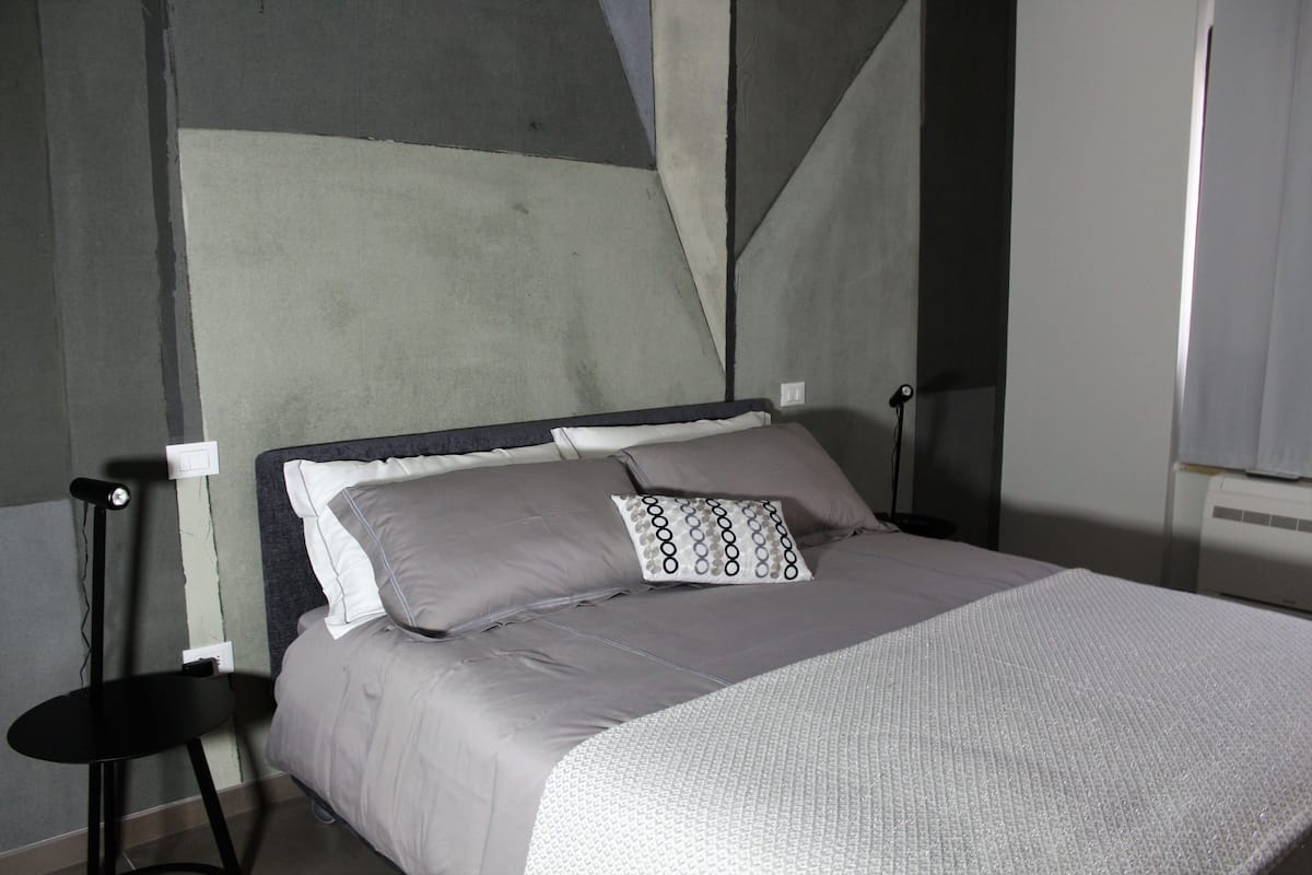 A neatly made bed with a grey and white patterned blanket, a pillow with a circular design, and a black side table with a lamp.