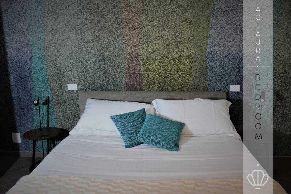 A bedroom with a textured wall, a bed with white linens and two teal pillows, and a small round table with a lamp.