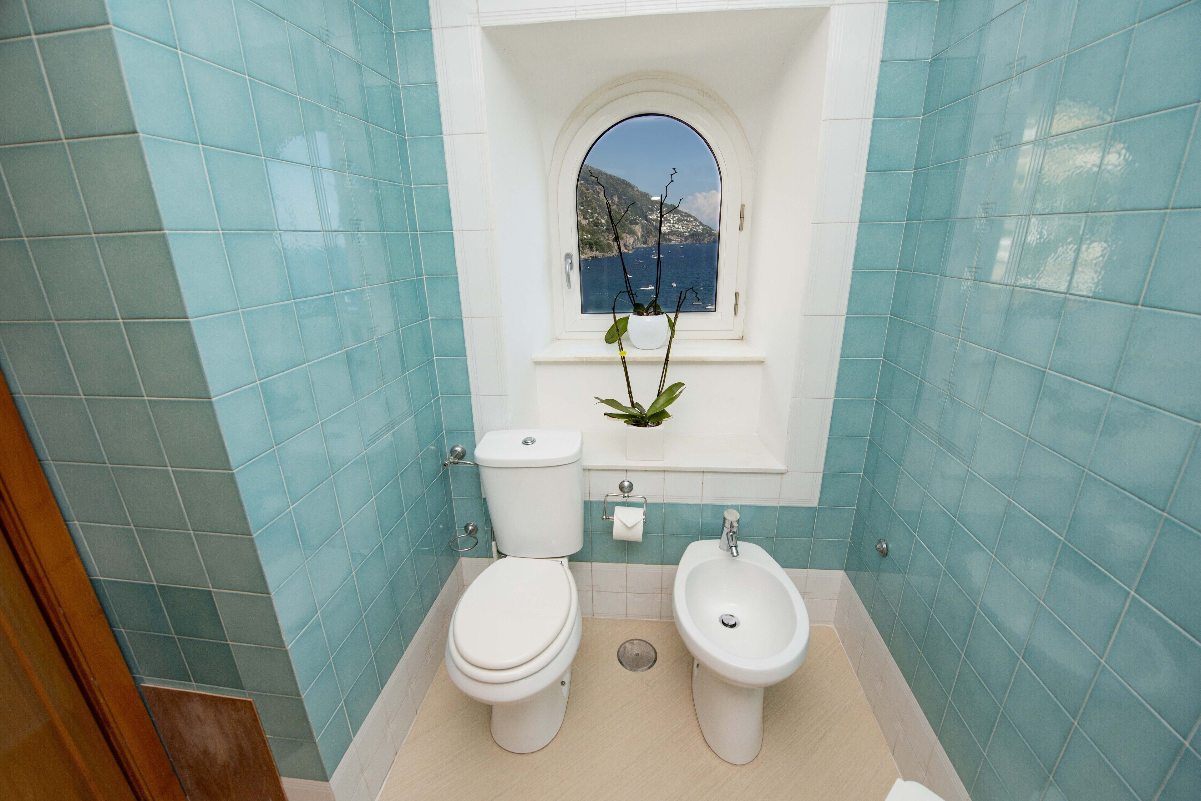 A bathroom with a toilet, bidet, and a window with a view of the sea.