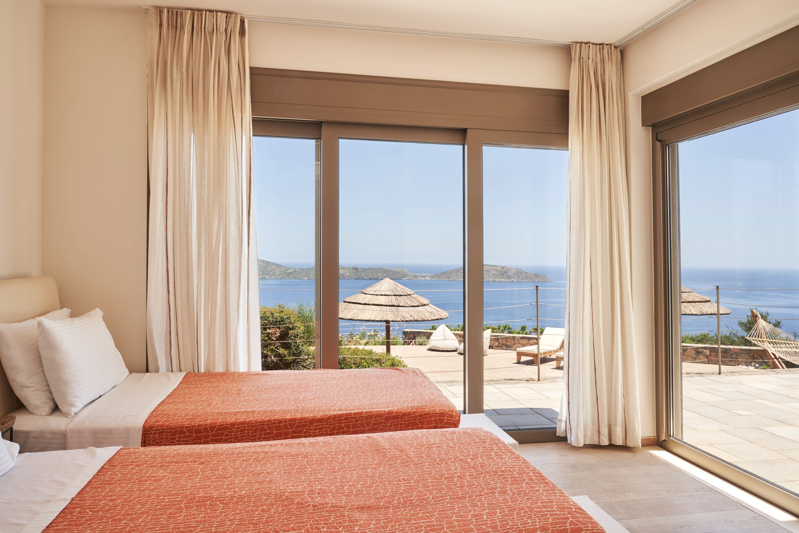 Habitación de hotel con dos camas, un ventanal con vista al mar y un área exterior con sombrilla de paja y sillones de descanso.