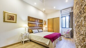 Una camera da letto con una testiera in legno, un letto con copripiumino bianco e rosa, un comodino con una lampada, una sedia in vimini e un muro in pietra.