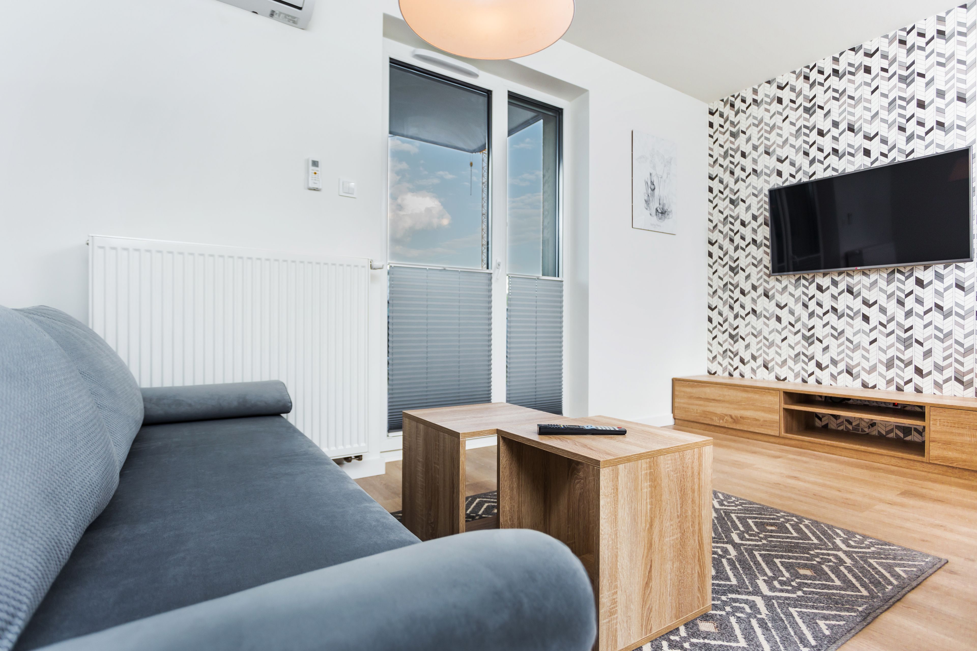 A modern living room with a grey sofa, a wooden coffee table, a TV on a patterned wall, and a window with blinds.