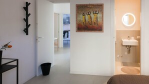 A modern hotel room with a bed, a bedside table, a black coat rack, a painting, a bathroom with a sink and mirror, and a hallway leading to another room.