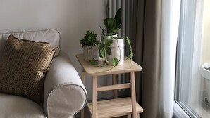 A living room with a beige sofa, a small wooden side table with plants, and a window with curtains. - In Matera "Sotto i pini" - COMFORT AND NEAR THE SASSI (Matera)