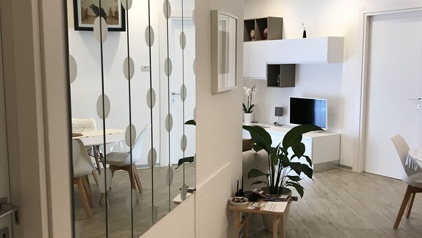 A modern living room with a large mirror, a dining area with chairs, a small table with a plant, and a TV on a white cabinet. - In Matera "Sotto i pini" - COMFORT AND NEAR THE SASSI (Matera)