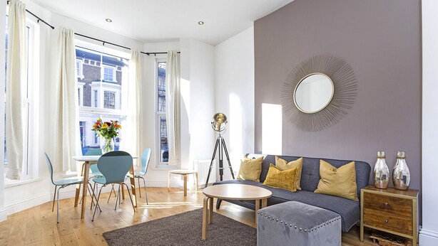 A modern living room with a sofa, ottoman, and a round coffee table. There is a dining area with chairs and a table, and a large window with curtains. - Luxury 2 Bed apartment- Zone 2 ( 10 mins into Central London)  (London)