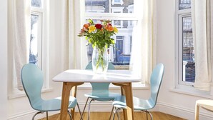 A dining area with a white table and two light blue chairs, a vase with flowers, and a view of buildings through large windows. - Luxury 2 Bed apartment- Zone 2 ( 10 mins into Central London)  (London)