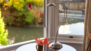 A window with a view of a river and greenery, a metal lamp, and a small vase with flowers on a table.