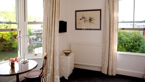 A room with a glass table, a chair, a bedside table with a lamp, a framed picture on the wall, and a window with curtains.