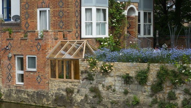 A brick building with a conservatory, a stone wall with plants, and a river.