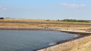 A coastal area with a sandy beach, a calm body of water, and wind turbines in the distance.