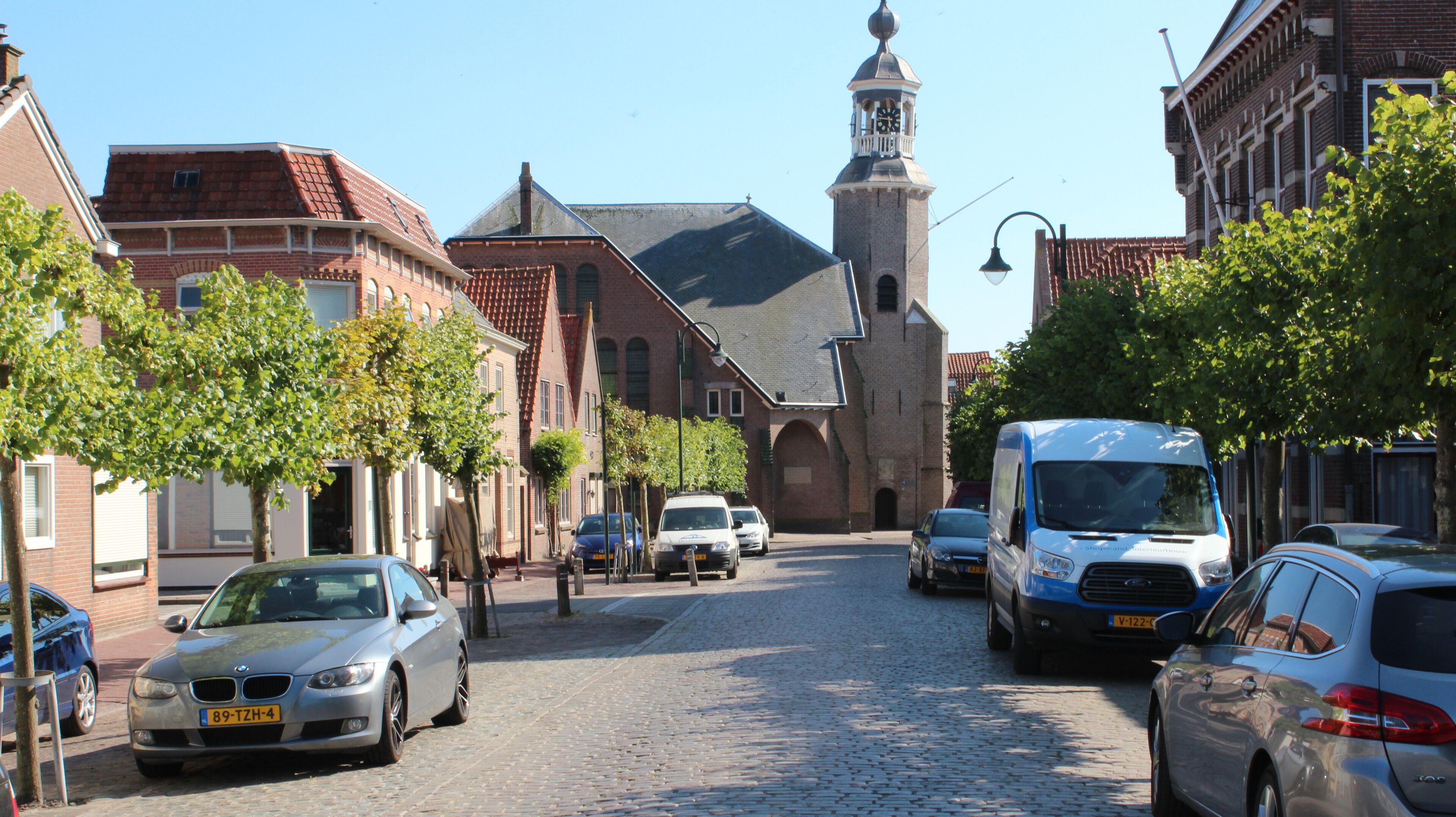 A cobblestone street lined with historic buildings, a church with a clock tower, and parked cars.