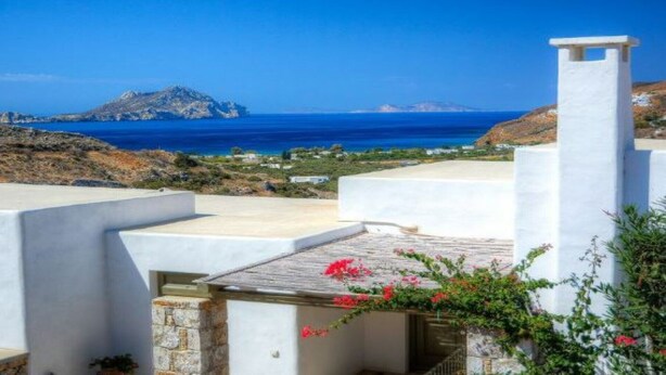 A white building with a stone base, a small garden with red flowers, and a view of the sea and a distant island.