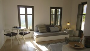 A cozy room with a bed, a small table, and a chair. There are two windows with shutters, a lamp on a side table, and a view of the outdoors.