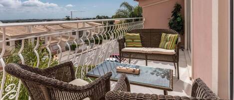 A balcony with wicker furniture and a glass table, overlooking a coastal area.