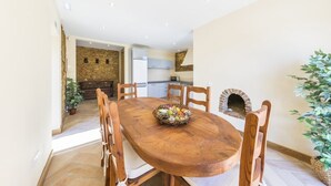 A dining area with a wooden table, chairs, and a central fruit basket. There is a stone wall and a kitchen in the background. - Villas Marim Flamingo Villa (Olhão)
