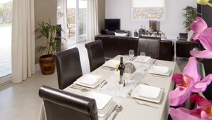Comedor con una larga mesa dispuesta para seis personas, rodeada de sillas de cuero oscuro. A la izquierda, una planta en maceta y a la derecha, un amplio ventanal con cortinas.