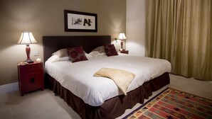 Una cama bien hecha con edredón blanco y marrón, dos mesitas de noche con lámparas, un cuadro en la pared y un ventanal con cortinas.