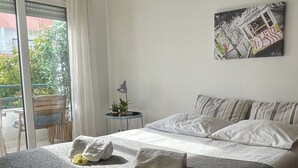 A bedroom with a bed, pillows, a bedside table with a lamp, a small potted plant, and a painting on the wall.