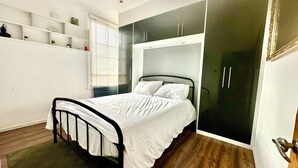 A bedroom with a bed, a wardrobe, a shelf unit, and a window with blinds. - Bright Brixton Apartment | Quick Access to Central London (London)