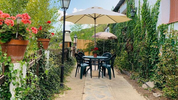 Un'area patio esterna con un vialetto in pietra, sedie di plastica verde, un tavolo e due ombrelloni che offrono ombra.