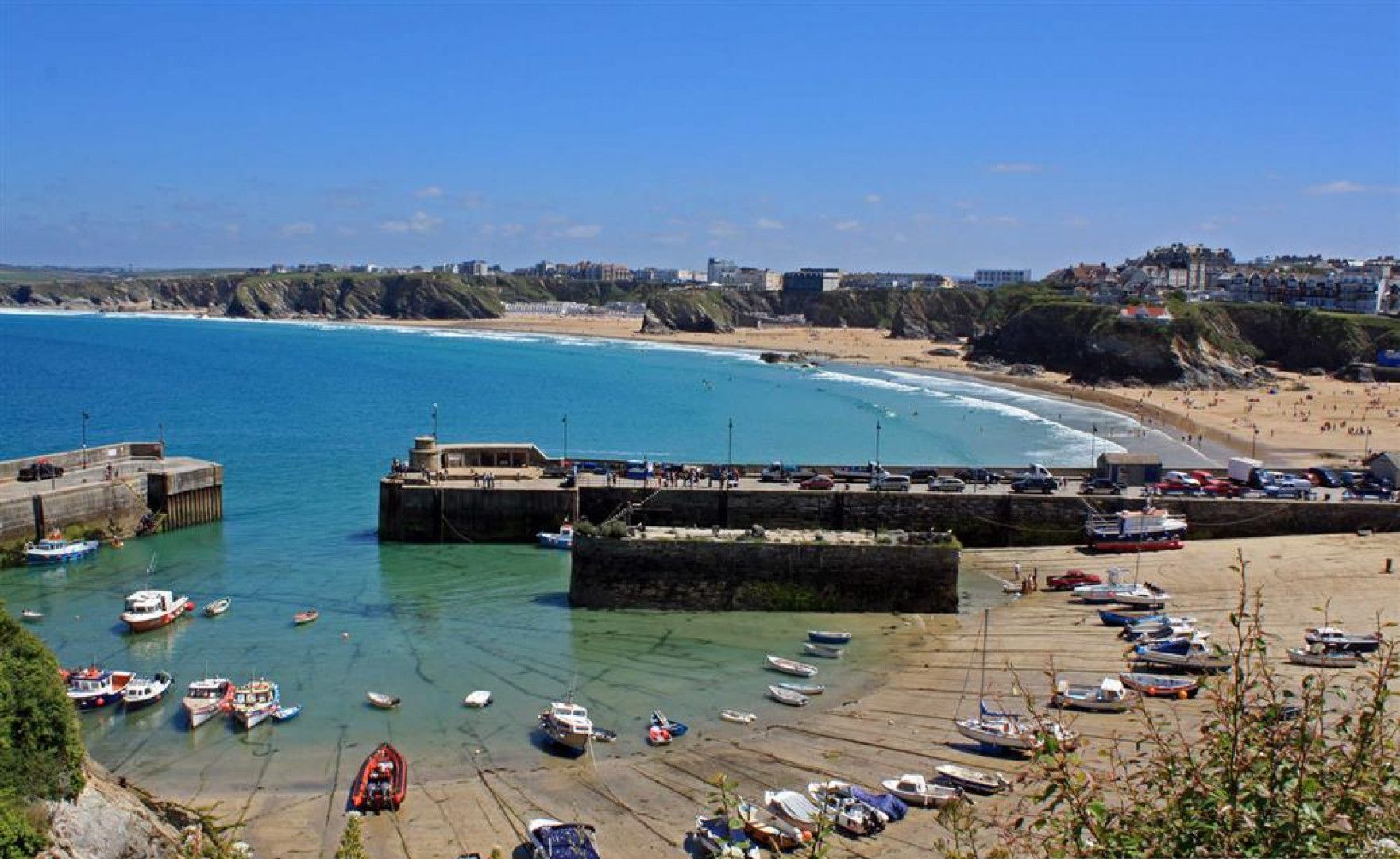 Un pueblo costero con un puerto deportivo, playa de arena y un acantilado rocoso.