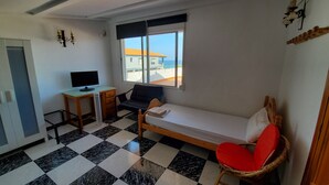 Una habitación con cama, escritorio con computadora, silla y ventana con vista a edificios y al mar.