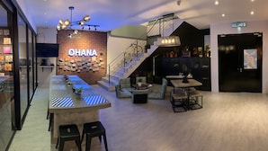 Un hall d’hôtel moderne comprenant un bar, des sièges, un escalier et une enseigne sur laquelle on peut lire « OHANA ».