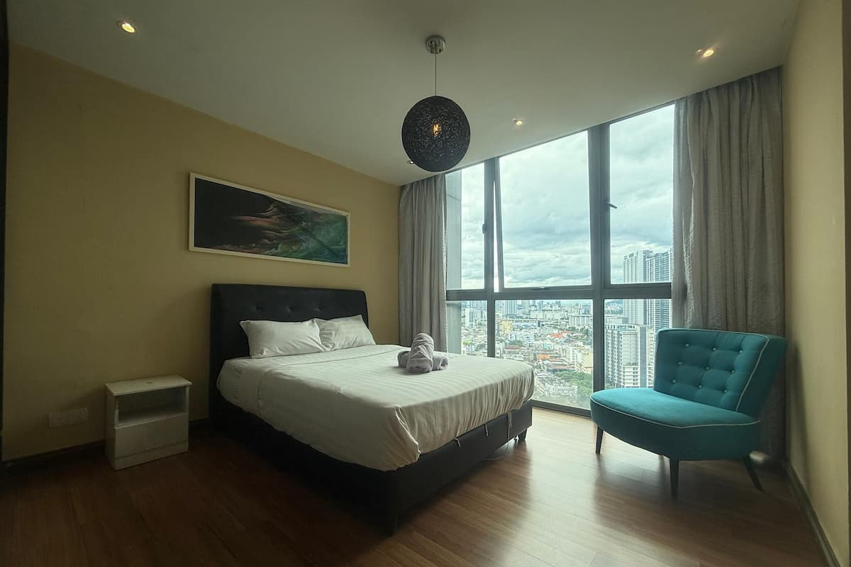 Une chambre moderne avec un grand lit, un fauteuil bleu et une vue sur la ville grâce à de grandes fenêtres.
