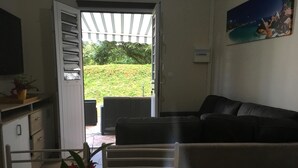 A living room with a sofa, a dining table with a vase of flowers, and a view of a garden through an open door. - Beautiful T3 in tropical forest with swimming pool (saint joseph)