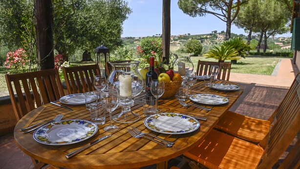 A round wooden table set for dining outdoors with chairs, plates, cutlery, glasses, and a basket of fruit. - Tenuta Santa Cristina - Luxury 8 beds Villa w/ private Pool and Tennis near Rome (Lazio)