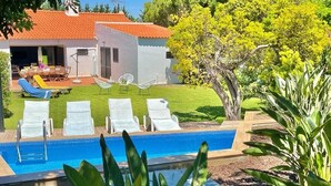 Zona de piscina amb gandules, una taula de dinar i un edifici amb teulada enrajolada.
