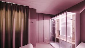 Una habitación de hotel moderna con ducha de vidrio, un ventanal con cortinas y una cama con cabecero.