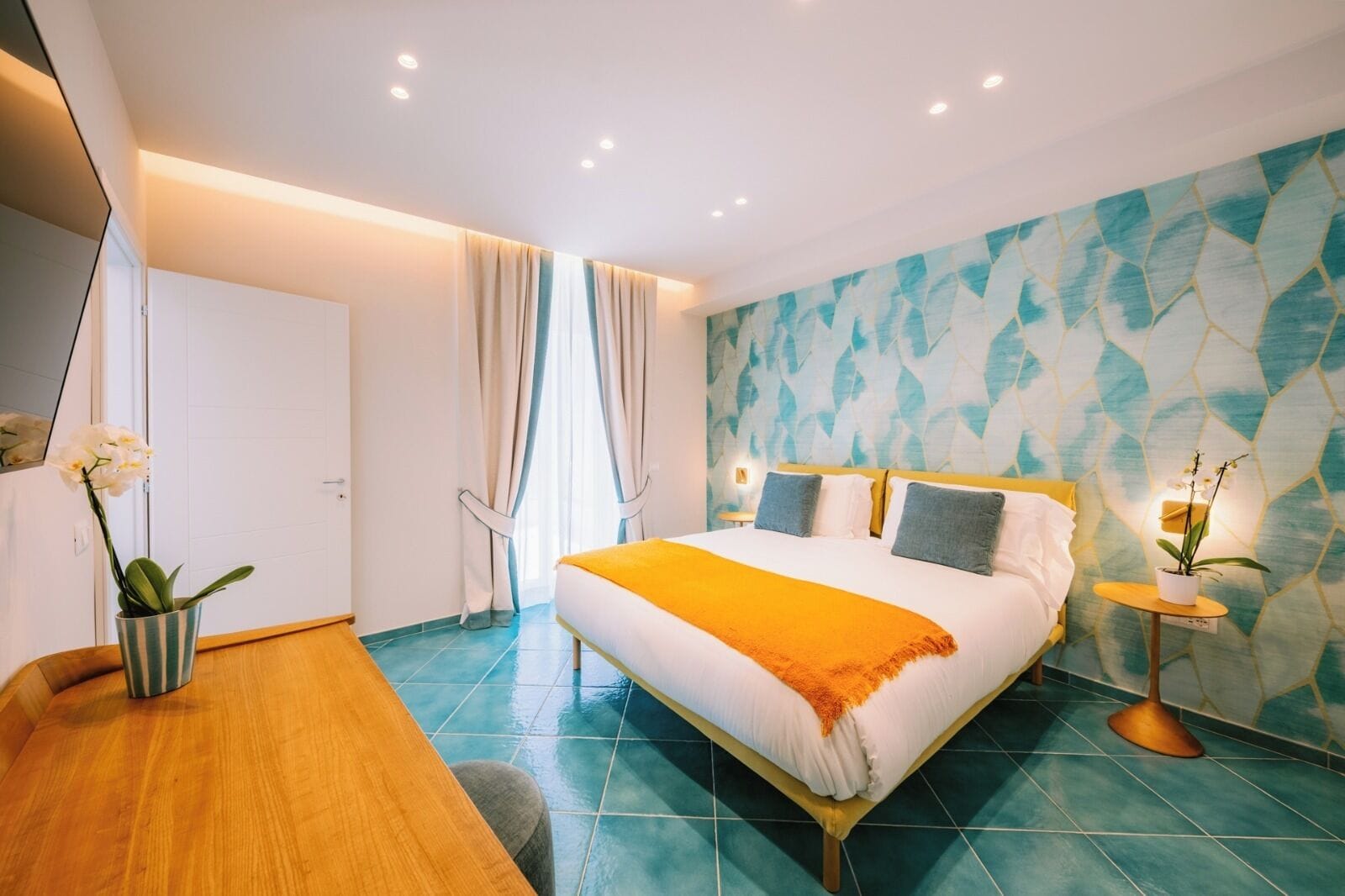 Quarto de hotel moderno com uma cama grande, uma mesinha de madeira, uma pequena mesa redonda com uma planta e um banheiro com porta branca.