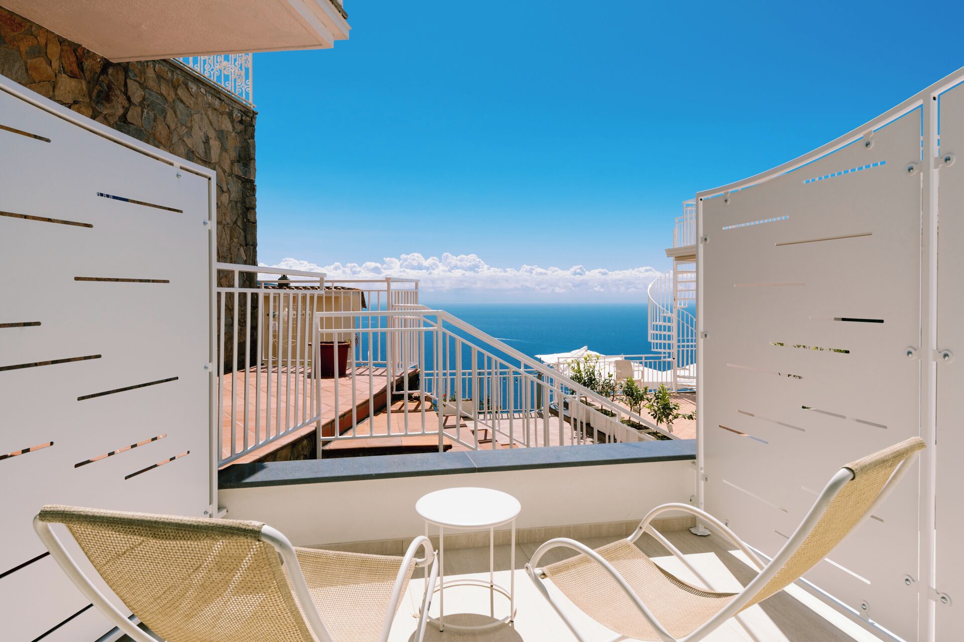 Un balcon avec des fauteuils en osier et une petite table, offrant une vue sur la côte, avec des bâtiments et un ciel dégagé.