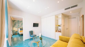 Una sala de estar moderna con un sofá amarillo, una mesa de centro de vidrio y un suelo de baldosas azules. Hay un televisor montado en la pared, una silla y una mesita con un jarrón de flores.