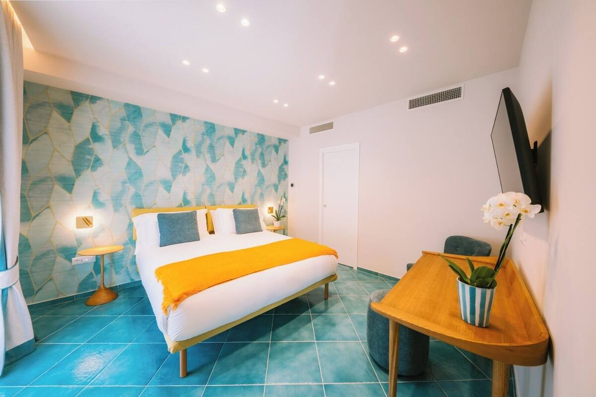 Una camera d'albergo con un letto, un comodino con una pianta, un tavolino e una TV a parete.