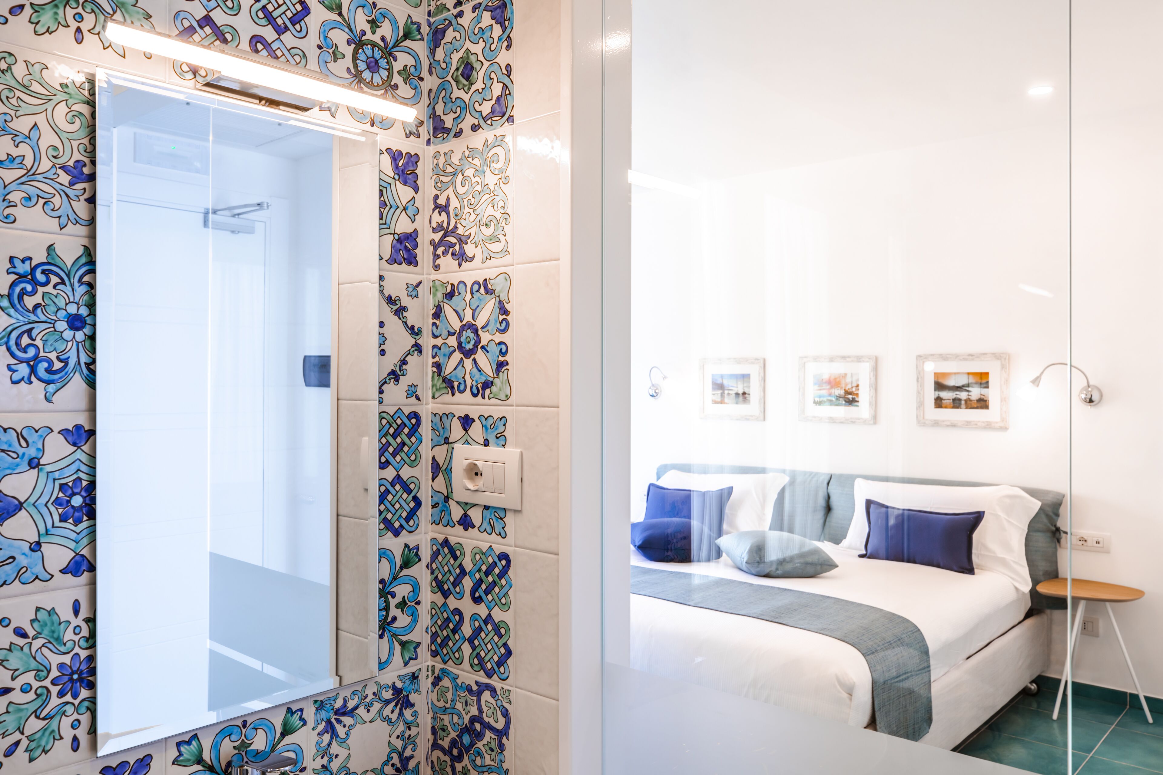 Un baño con ducha y espejo, una cama con ropa de cama azul y blanca, y una pared con cuadros enmarcados.