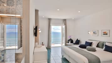 A modern hotel room with a large bed, a balcony with a view of the sea, and a small table with a chair.