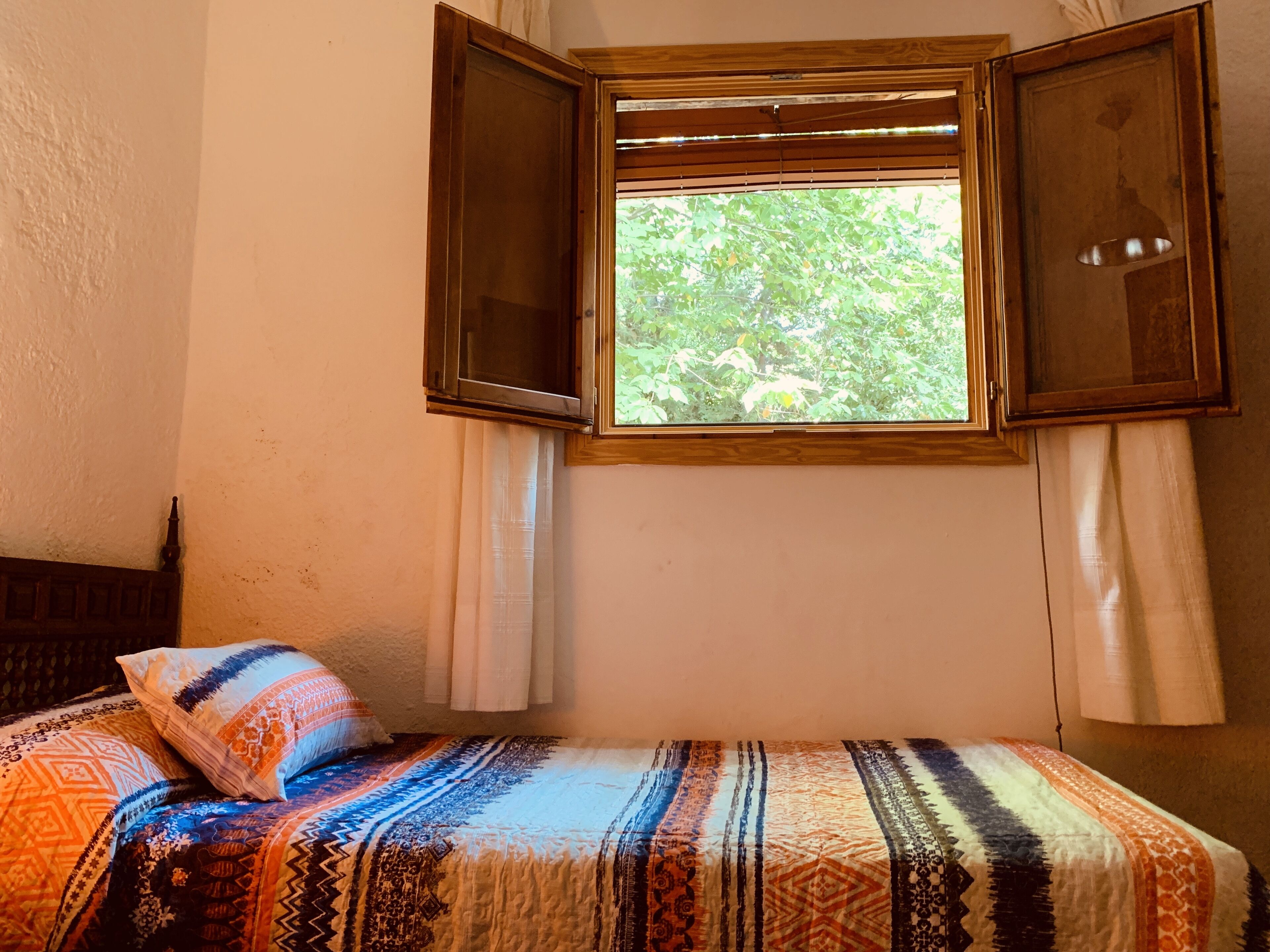 A room with a bed, a window with open shutters, and a view of greenery outside.