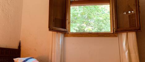 A room with a bed, a window with open shutters, and a view of greenery outside.