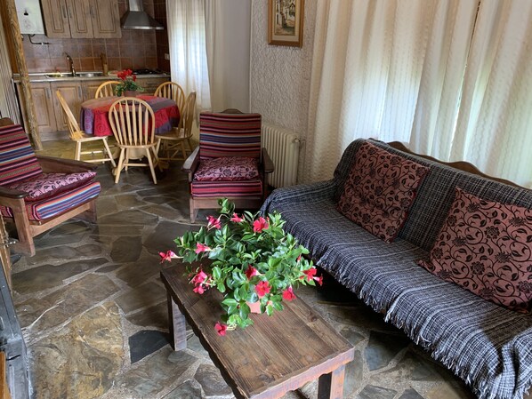 Un salón acogedor con suelo de piedra, muebles de madera, un sofá con cojines estampados y una mesa con un arreglo floral.