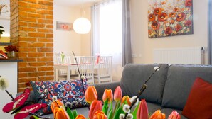 Een woonkamer met een bakstenen muur, een grijze bank, kleurrijke kussens en een schilderij van bloemen aan de muur.