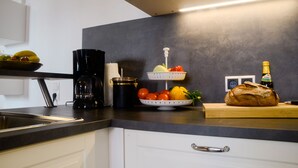 Een keuken met een koffiezetapparaat, een schaal met fruit, een mand met groenten en een brood op het aanrecht.
