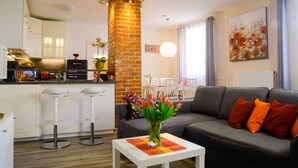 Een moderne woonkamer met een grijze bank, een salontafel met een bloevas, en een keuken met witte kasten en een bakstenen zuil.