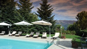 Area piscina con lettini e ombrelloni, circondata da alberi con montagne sullo sfondo.