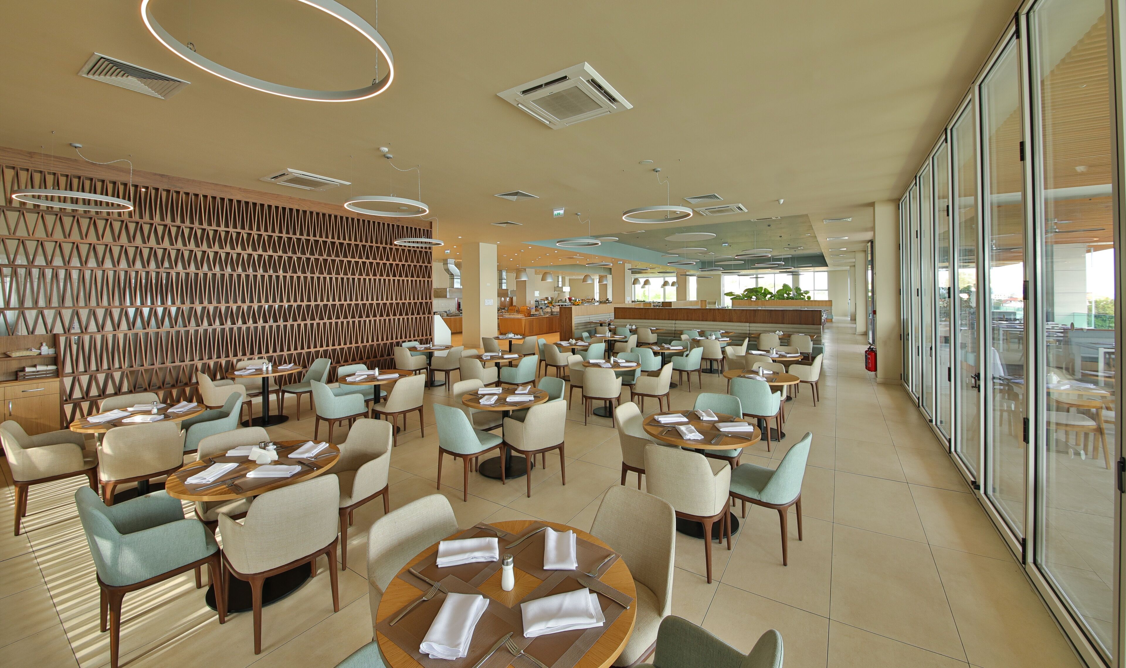 A spacious restaurant with round tables and chairs, a modern ceiling design, and a large glass wall offering a view.