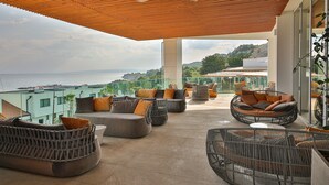 A spacious outdoor seating area with wicker furniture and cushions, overlooking a scenic view of the sea and hills.