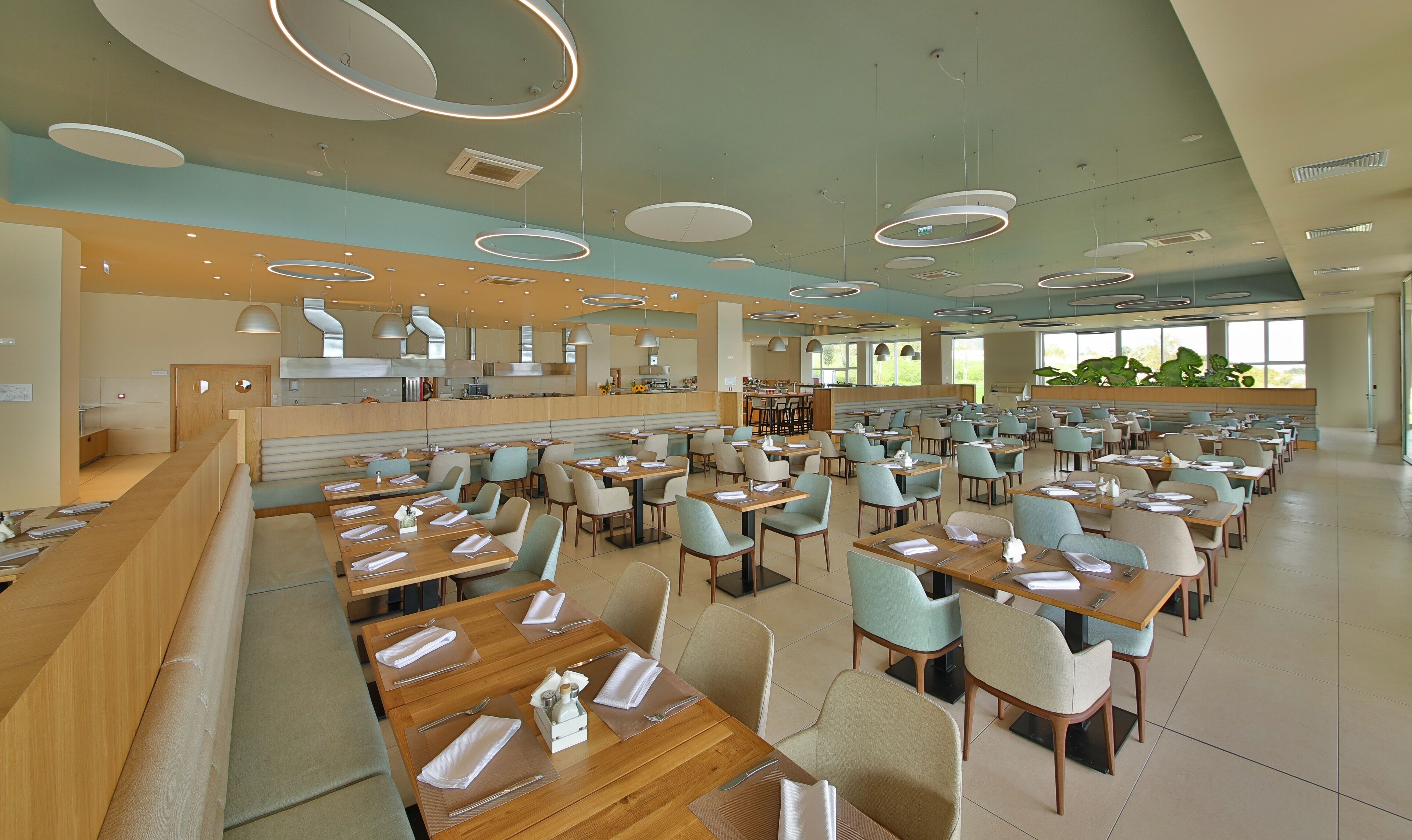 A spacious dining area with multiple tables and chairs, a kitchen in the background, and large windows providing natural light.